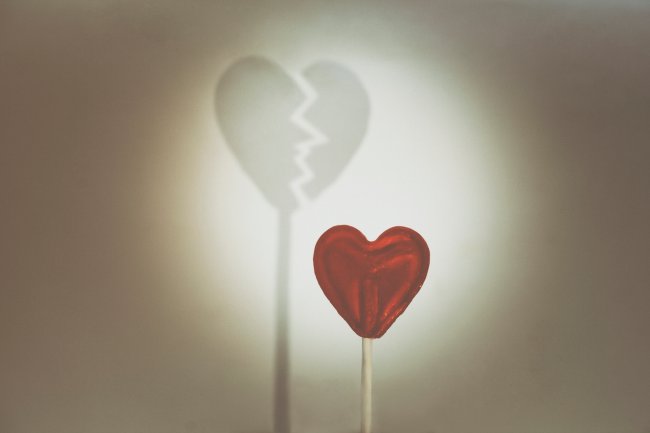 Ilustração de um pirulito vermelho de coração e, atrás dele, uma sombra de um coração partido