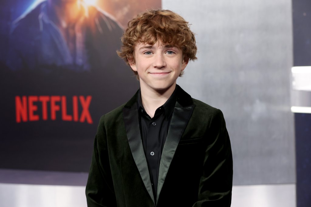 Walker Scobell na premiere de The Adam Project; ele está de terno preto sorrindo levemente enquanto posa para foto com pôster do filme da Netflix ao fundo