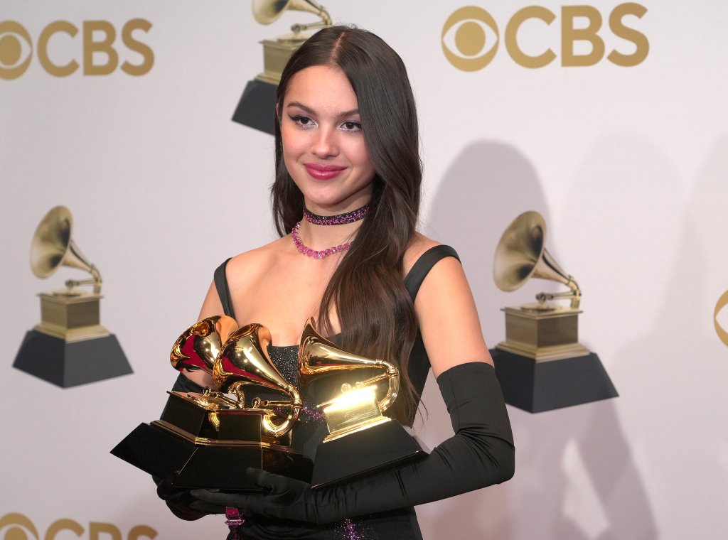 Olivia Rodrigo com seus prêmios no GRAMMYs; ela está segurando três troféús e sorrindo levemente com o backdrop da premiação ao fundo