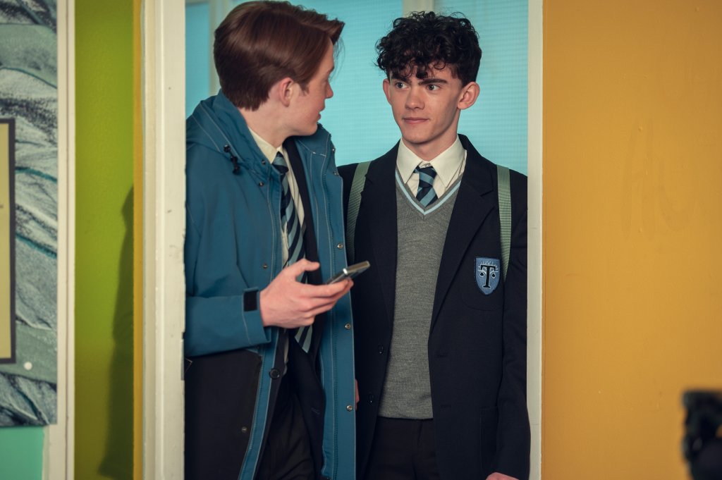 Nick e Charlie, de Heartstopper; eles estão se olhando enquanto passam por uma porta da escola