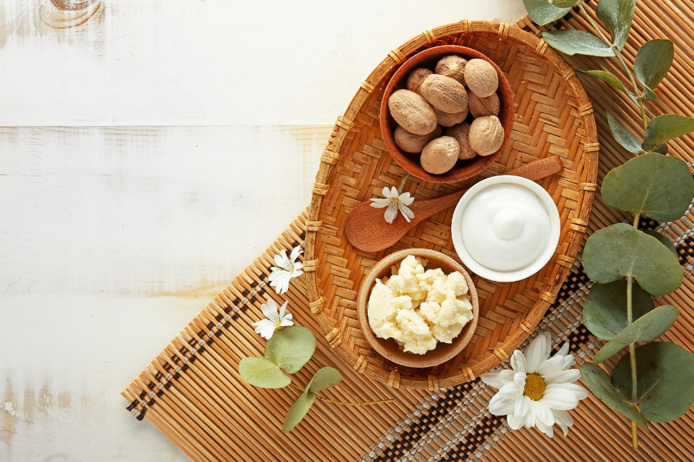 Imagem mostra uma tábua de madeira repleta de frutos do karité, manteiga de karité, folhas e flores.