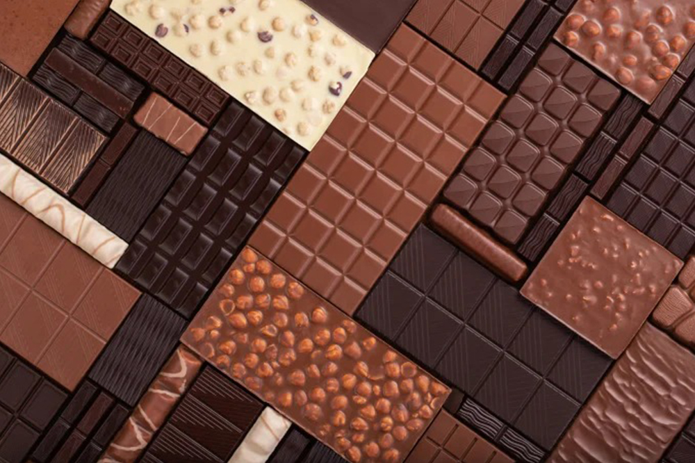 Imagem mostra tabletes de chocolate de vários tipos: ao leito, branco, amargo, crocante. Todos posicionados assimetricamente, um do lado do outro.
