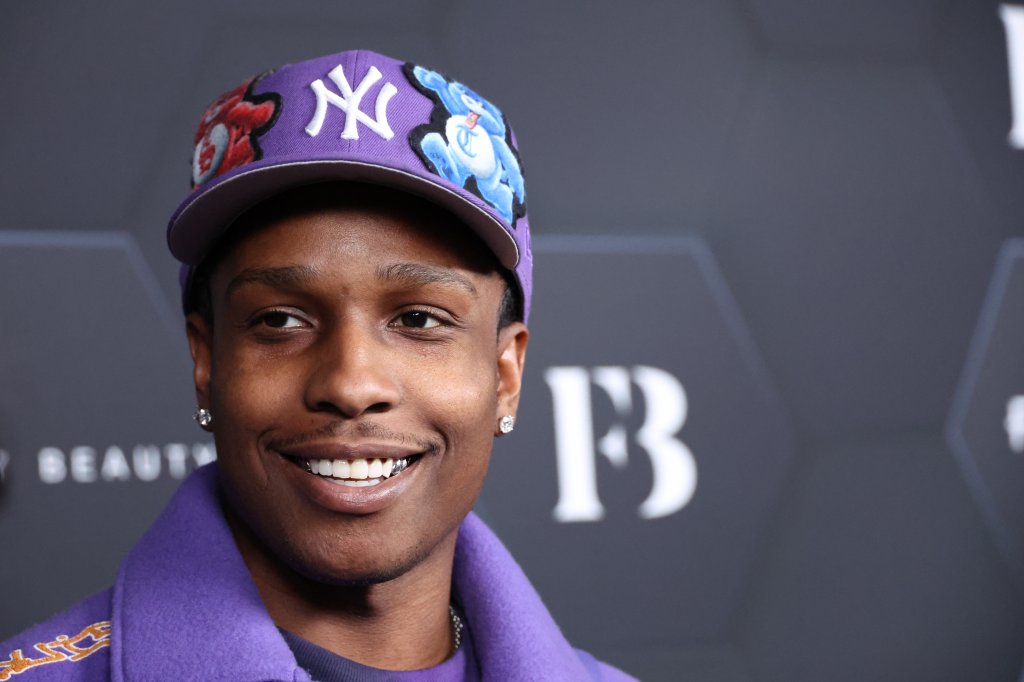 A$AP Rocky sorrindo para foto com o backdrop da Fenty Beauty ao fundo; ele usa boné e casaco na cor lilás