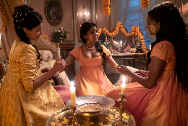 bridgerton-cerimonia-casamento-edwina Foto de uma cena da segunda temporada de Bridgerton. Na imagem está a família Sharma, na parte esquerda está Mary Sharma com um vestido amarelo e cabelo preso em um coque, no meio Edwina Sharma com um vestido laranja e cabelo solto em uma trança e na parte direita Kate Sharma com um vestido laranja e cabelo meio preso. As três estão sentadas no chão fazendo a Cerimônia Haldi para o casamento.