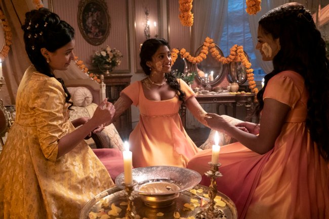 Foto de uma cena da segunda temporada de Bridgerton. Na imagem está a família Sharma, na parte esquerda está Mary Sharma com um vestido amarelo e cabelo preso em um coque, no meio Edwina Sharma com um vestido laranja e cabelo solto em uma trança e na parte direita Kate Sharma com um vestido laranja e cabelo meio preso. As três estão sentadas no chão fazendo a Cerimônia Haldi para o casamento.