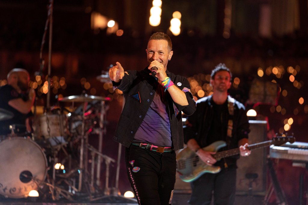 Chris Martin sorrindo enquanto canta em apresentação do Coldplay; ele aponta com uma das mãos e segura o microfone com a outra