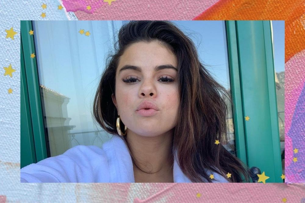 Montagem com o fundo colorido e detalhe de estrelas coloridas com a foto da Selena Gomez no centro. A foto é uma selfie e ela usa uma camisa branca, brincos de argola dourados, olha para a câmera e faz um bico.