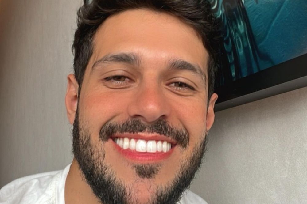 Selfie de Rodrigo Mussi sorrindo; ele usa uma camiseta branca e está de barba