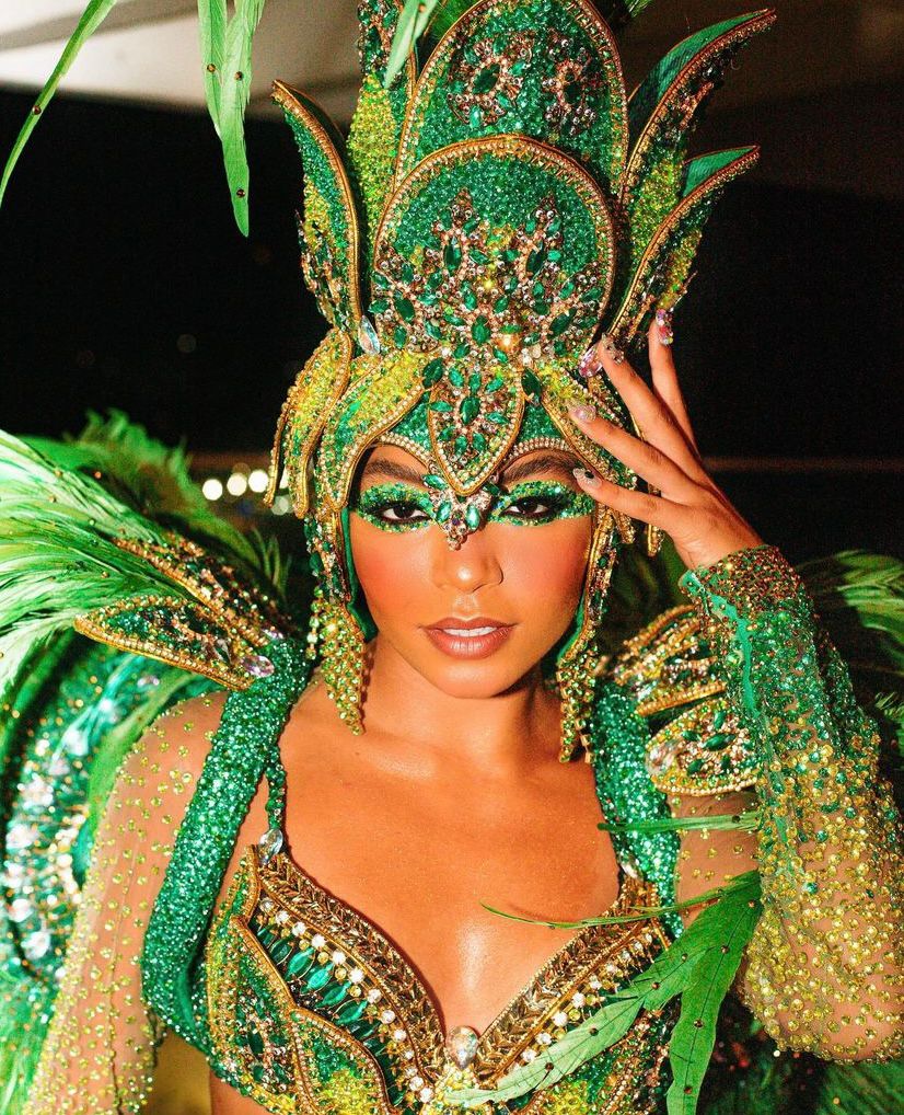 A imagem mostra em destaque Any Gabrielly maquiada e adornada para o desfile da escola de samba carioca Unidos de Vila Isabel, na Marquês de Sapucaí. A imagem segue acompanhada de uma borda colorida e estrelinhas douradas por toda a imagem.