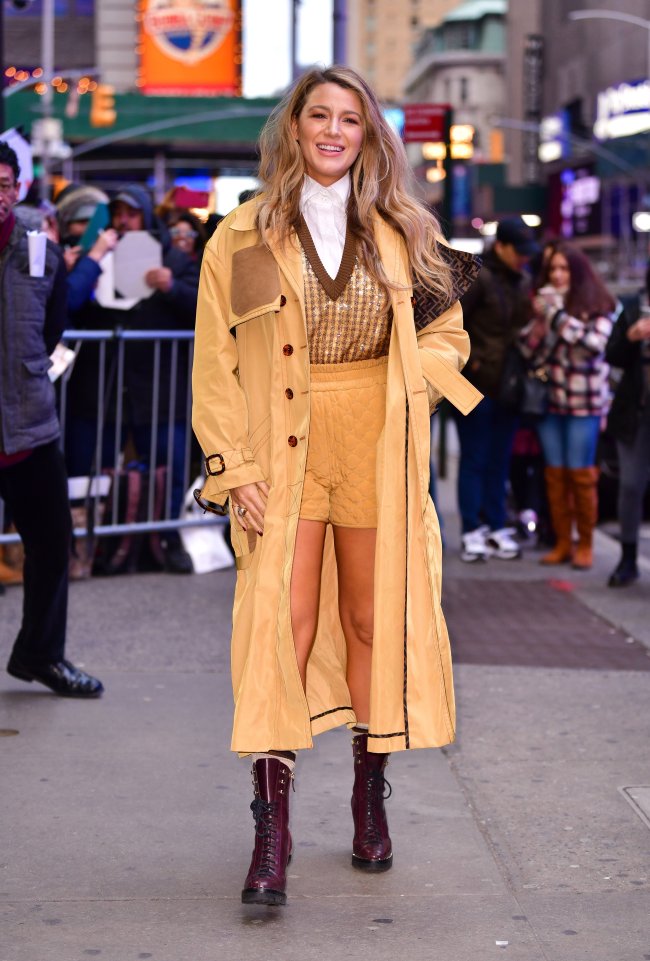 Foto Blake Lively com blusa marrom e bege, bermuda bege e casaco alongado bege. Ela está na rua e sorri.