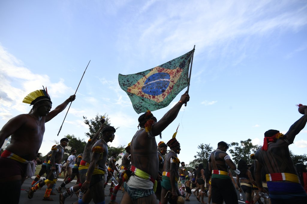 Protesto de povos indígenas contra o garimpo ilegal. Eles seguram cartazes e a bandeira do Brasil cobertos de tinta vermelha, que representa o sangue