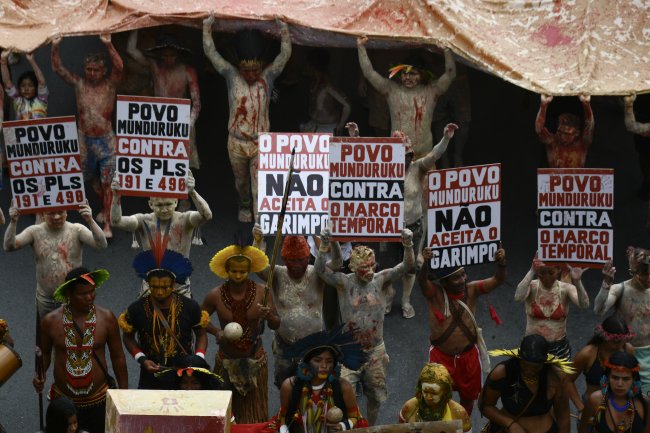 Protesto de povos indígenas contra o garimpo ilegal. Eles seguram cartazes e a bandeira do Brasil cobertos de tinta vermelha, que representa o sangue