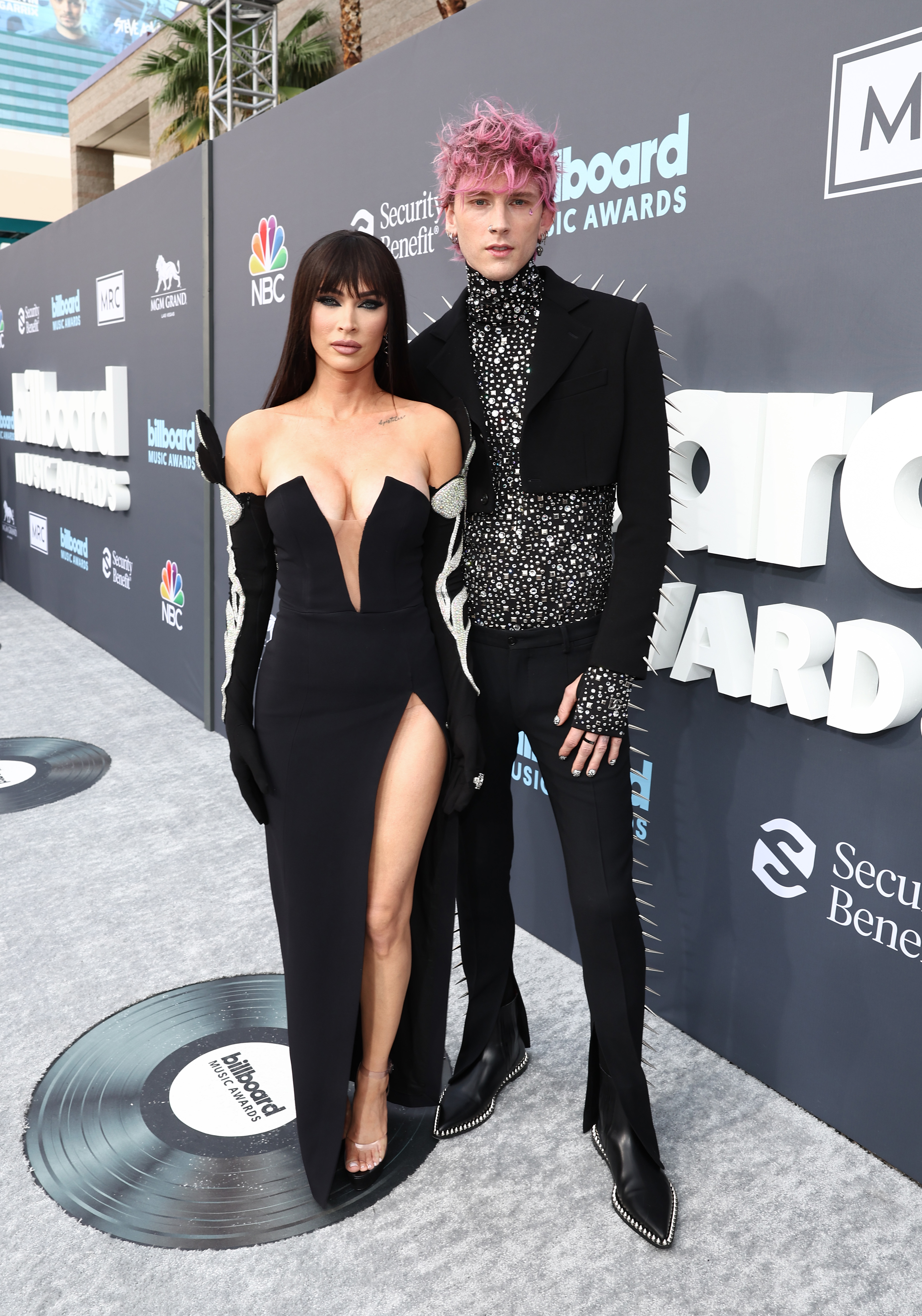 Megan Fox usando vestido preto com fenda lateral e luvas e Machine Gun Kelly todo de preto no Billboard Music Awards 2022