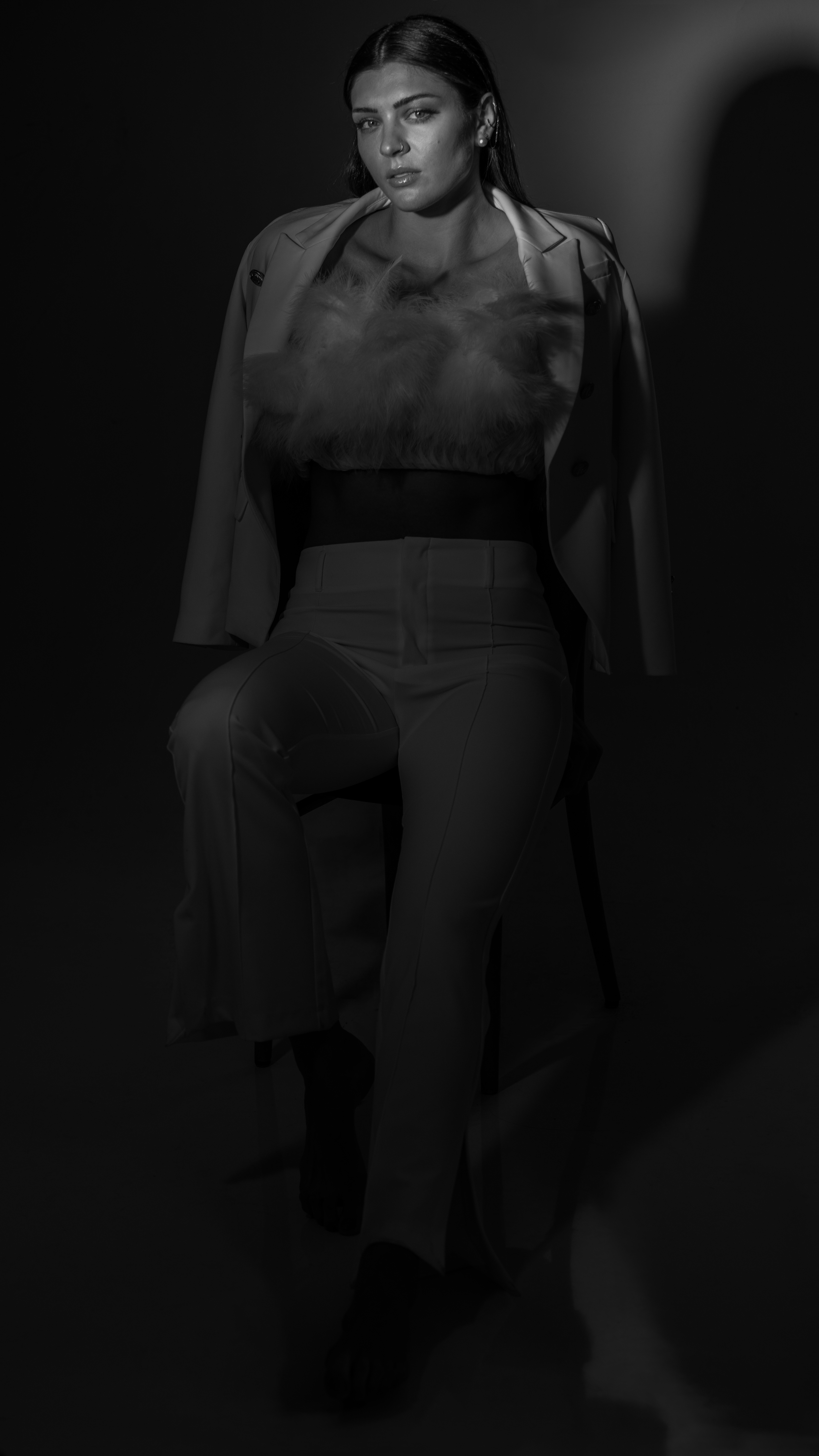 Imagem mostra Rosamaria Montibeller sentada em frente a um fundo infinito usando blazer branco e cropped tomara que caia com plumas, enquanto olha para câmera. A imagem tem uma tonalidade cinza e preta e uma pequena luz clara que destaca o rosto de Rosa, da direita para a esquerda.