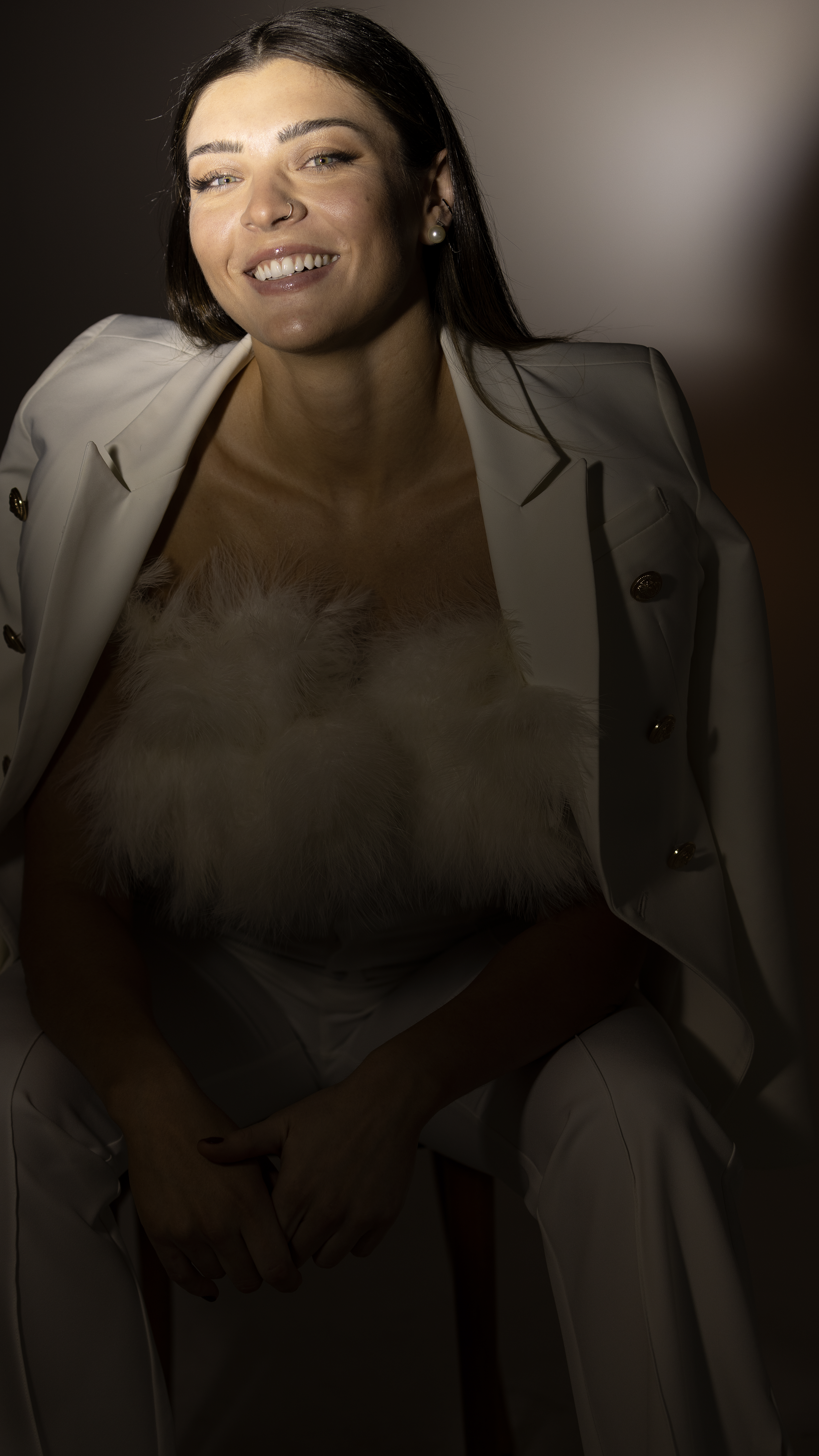 Imagem mostra Rosamaria Montibeller posando sentada em frente a um fundo infinito usando blazer branco e cropped tomara que caia com plumas, enquanto sorri para câmera. A imagem tem uma tonalidade cinza e preta e uma luz clara destacando o rosto de Rosa.