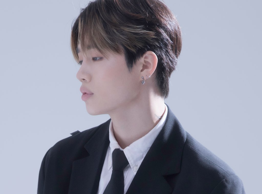 Foto de perfil de Spax; ele está em um fundo cinza com expressão neutra usando um terno com gravata preta e camisa branca