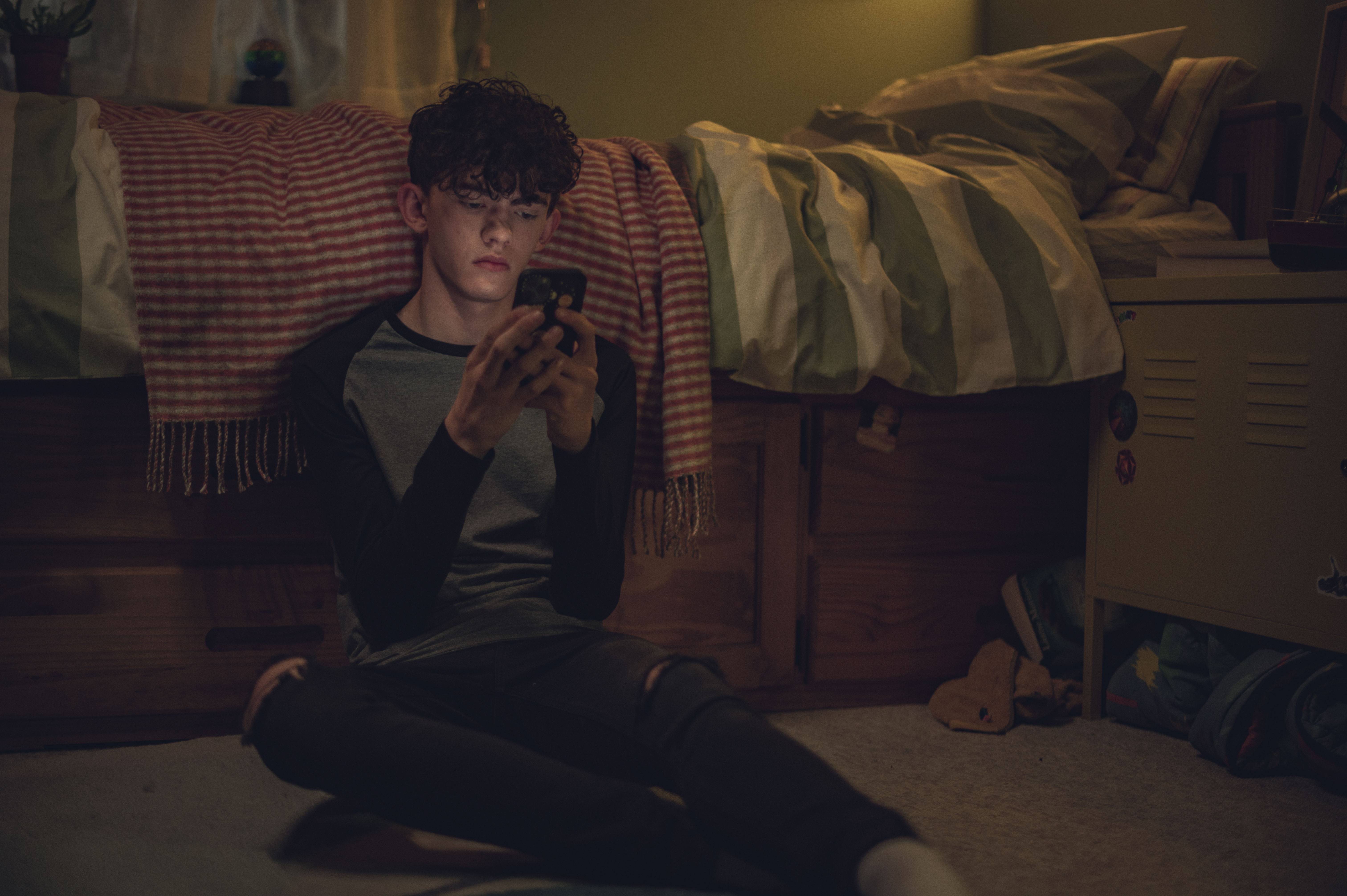 Charlie Spring (Joe Locke) em Heartstopper. Ele está sentado no chão ao lado de sua cama, digitando no celular, usando blusa de manga comprida e calça preta rasgada nos joelhos