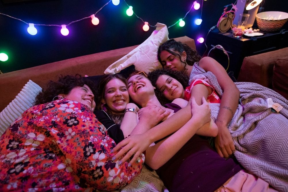 Várias meninas abraçadas em um sofá, com luzes coloridas no fundo