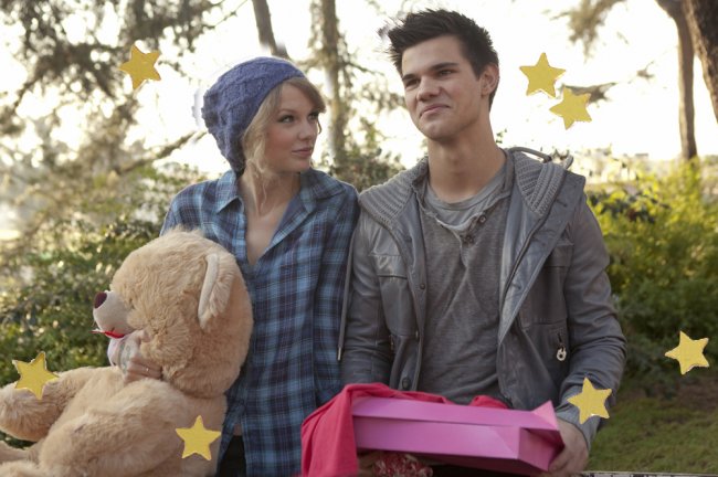 Os personagens de Taylor Swift e Taylor Lautner no filme Idas e Vindas do Amor. Taylor segura um enorme urso de pelúcia e Lautner uma caixa rosa de bombons