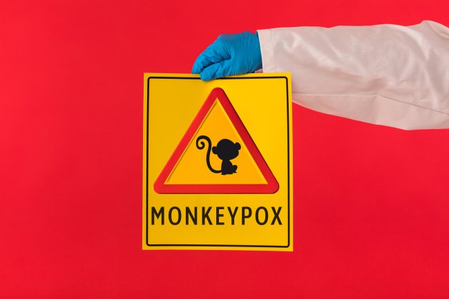 Uma mão com luvas azul segura uma placa sinalizando a varíola dos macacos; tem um macaquinho desenhado nela, que é amarela