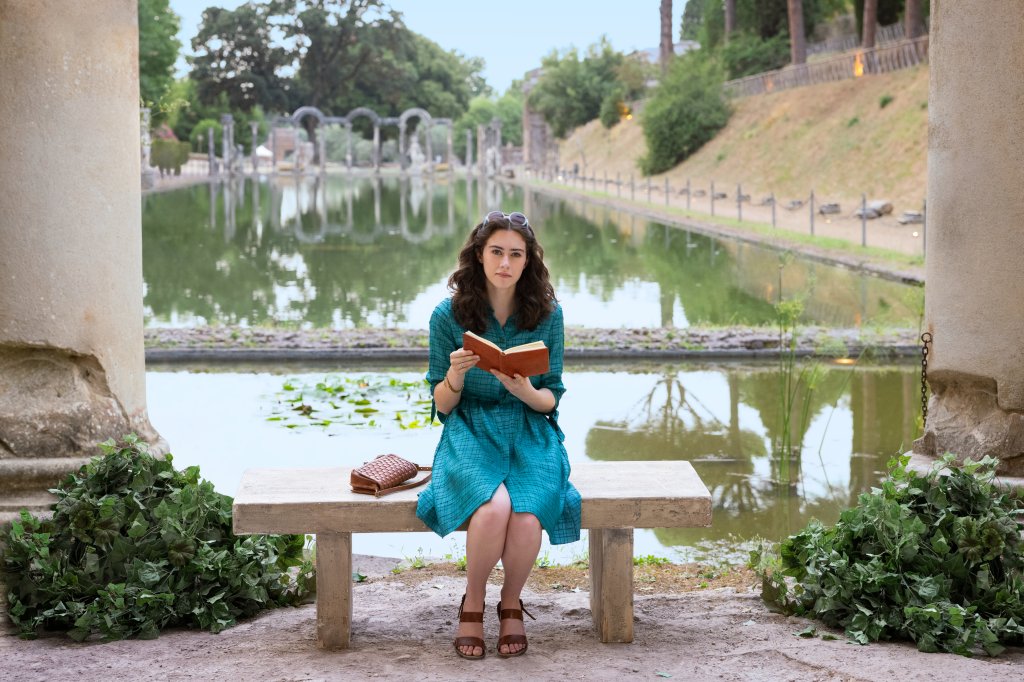 Susanna Skaggs como Lina; ela está sentada com um lago ao fundo com um livro nas mãos e olhando para frente; a personagem usa um vestido azul