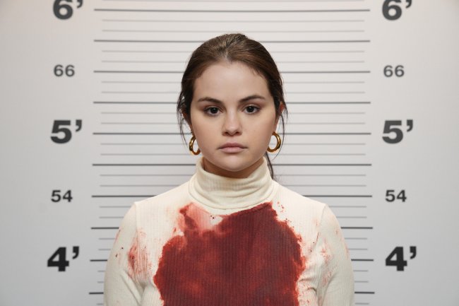 Selena Gomez caracterizada como Mabel em Only Murders in The Building; ela está com expressão neutra posando para foto da polícia com medidores de altura ao fundo e sua blusa está manchada de sangue
