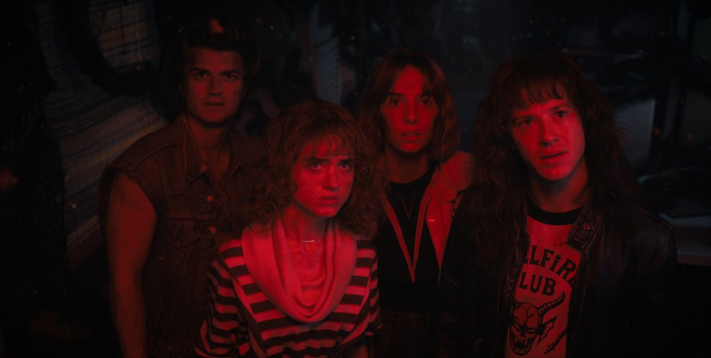 Steve, Nancy, Robin e Eddie em cena de Stranger Things; eles estão olhando para cima com expressões confusas e com os rostos iluminados por uma luz vermelha