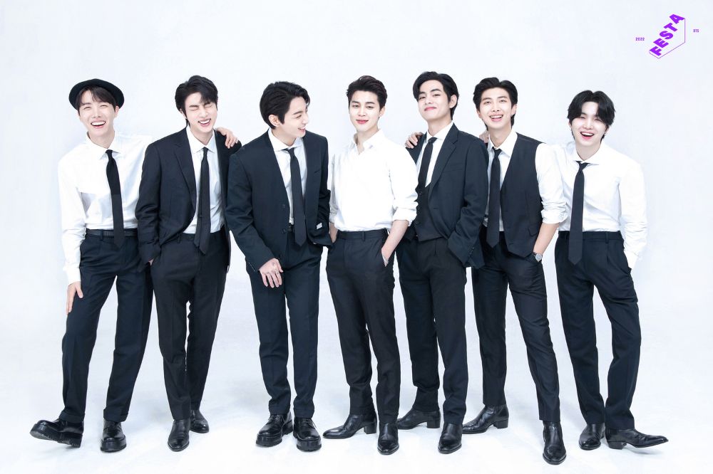 Integrantes do BTS alinhados na horizontal usando ternos pretos e sorrindo enquanto posam em um fundo branco com o logo roxo da BTS Festa 2022 no canto superior direito da imagem