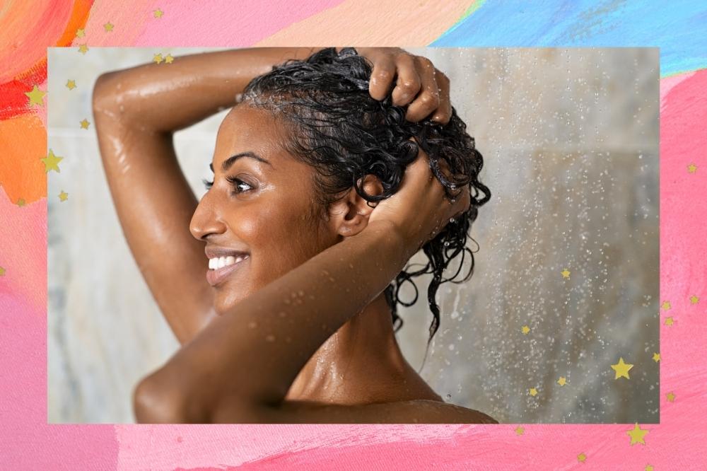Montagem com o fundo rosa e detalhe de estrelas douradas com a foto de uma mulher lavando o cabelo no centro. Ela está de perfil, com as mãos no cabelo e sorri.