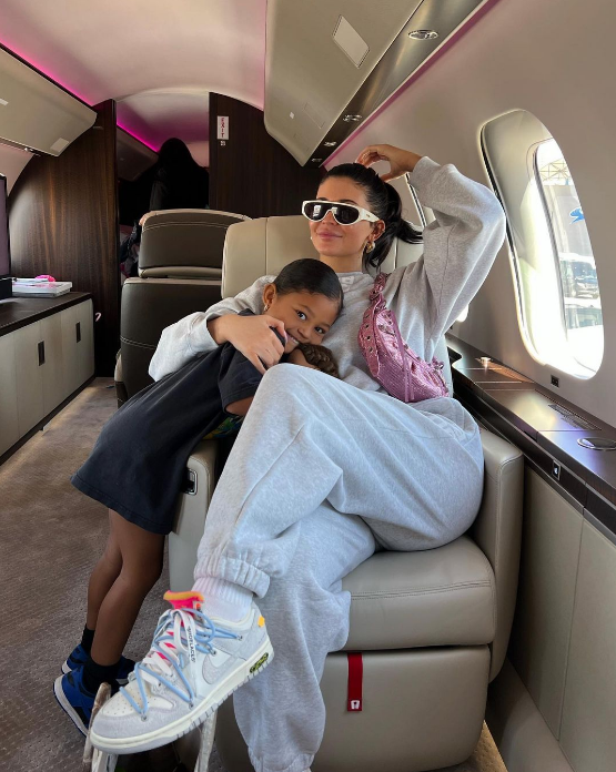 Kylie Jenner e Stormi Webster abraçadas em jatinho. Kylie está usando conjuntinho de moletom cinza e bolsa Le Cagole rosa, da Balenciaga