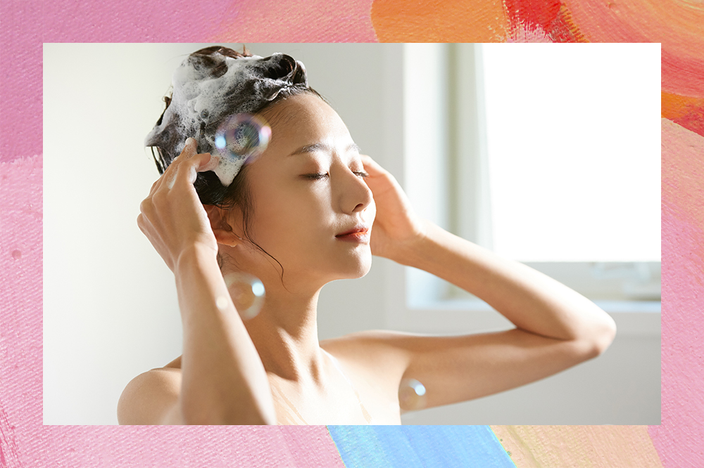 Menina lavando cabelo no banho com olhos fechados e as duas mãos na cabeça. O cabelo está com espuma de xampu