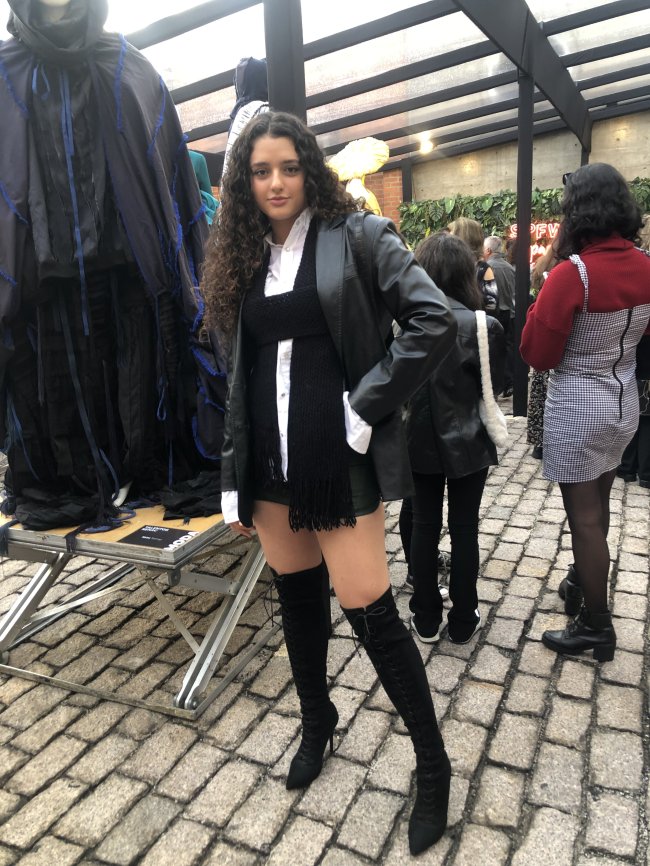 Foto da Giovanna Venturoli na São Paulo Fashion Week nº53 usando blazer de couro, camisa branca, saia preta e bota preta over the knee. Ela olha para a câmera e sorri.