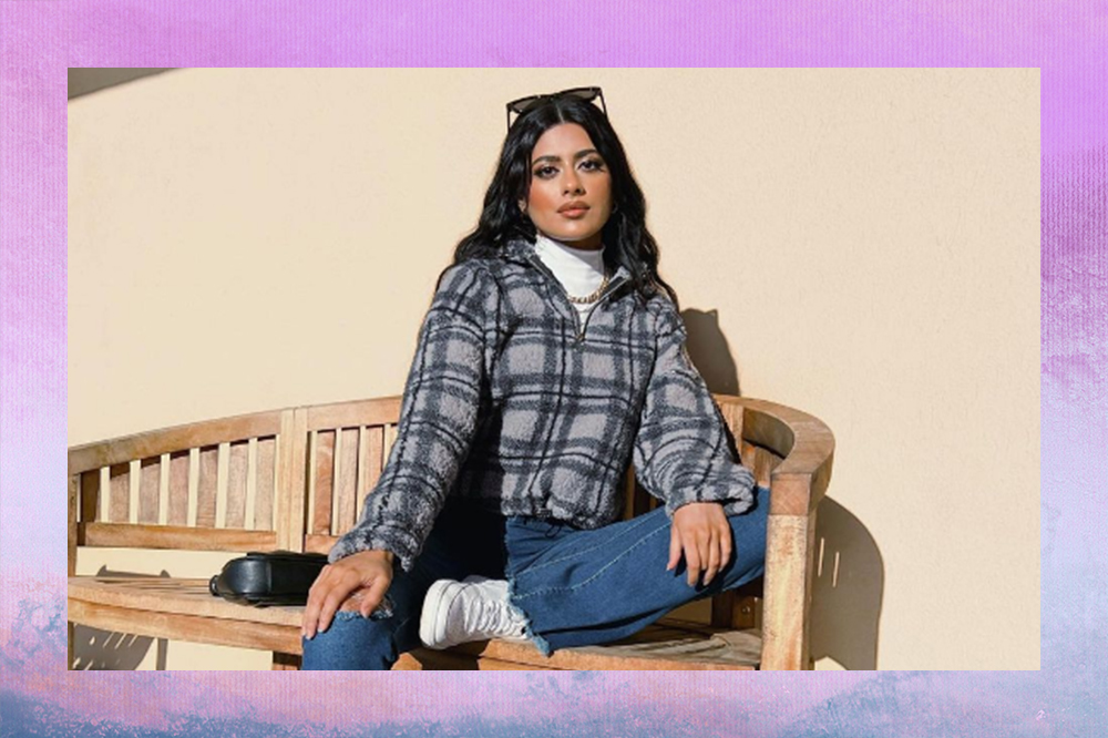 Foto de garota sentada em um banco usando blusa branca de gola alta por baixo de casaco xadrez, calça jeans e tênis branco. Ela está com um óculos de sol no cabelo e uma bolsa preta jogada no banco. O fundo da montagem é degradê em azul e roxo.