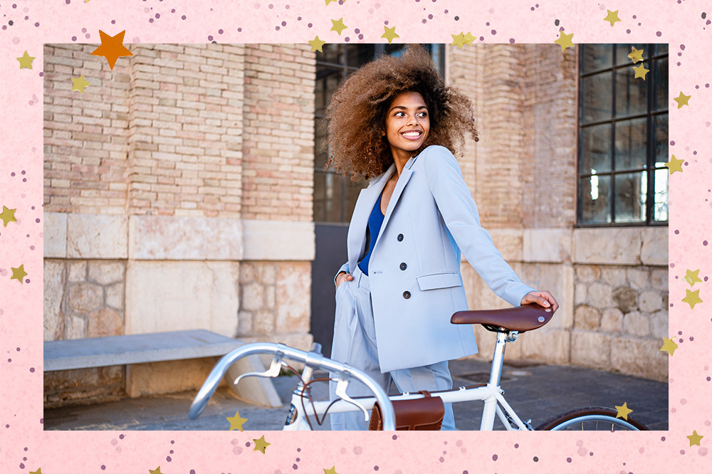 Montagem em fundo rosa com textura roxa com pinceladas de tinta e detalhes de estrelas douradas de foto de menina usando blazer e calça de alfaiataria em tonalidade azul pastel posando apoiada em uma bicicleta.
