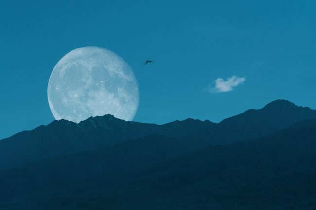 Lua Cheia nascendo por trás de uma montanha. Pássaros voam no céu