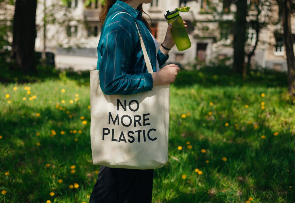 Uma mulher bebendo água direto de uma garrafa reutilizável e segurando uma ecobag que diz "não mais plástico"