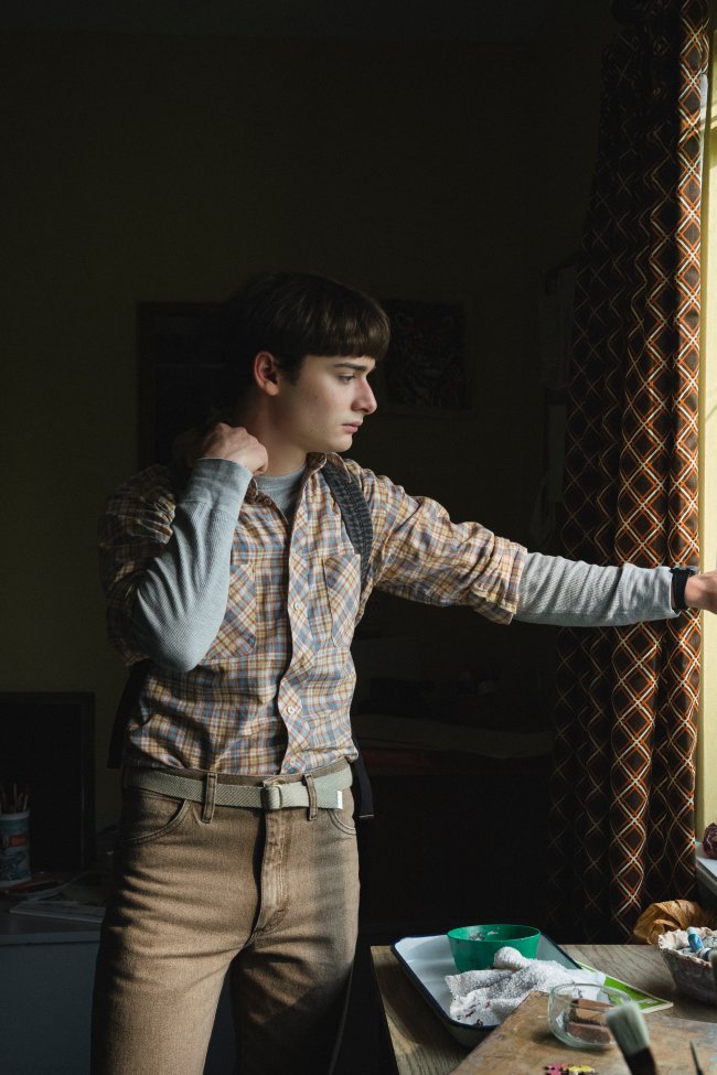 Noah Schnapp como Will Byers em Stranger Things; ele está com uma das mãos atrás do pescoço e a outra segurando uma cortina enquanto ele olha para fora da casa pela janela