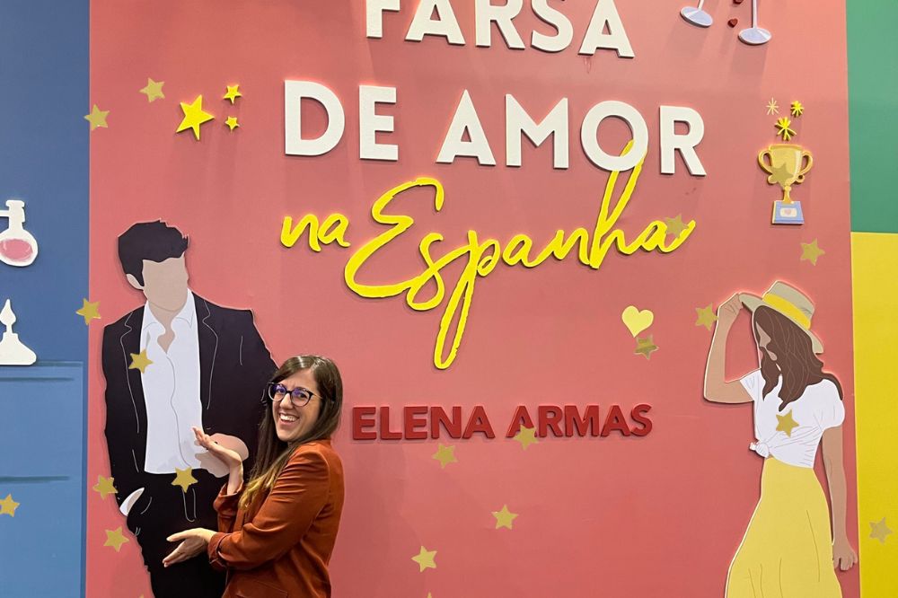 Elena Armas posando no estande da Arqueiro com a capa de seu livro "Uma Farsa de Amor na Espanha" ao fundo; ela está sorrindo próxima ao desenho do personagem Aaron; estrelas amarelas decoram a imagem