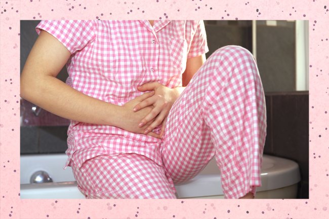 Jovem sentada em uma banheira branca coloca as duas mãos sobre a barriga, como se ela estivesse sentindo cólicas. Ela veste um pijama quadriculado rosa e branco.