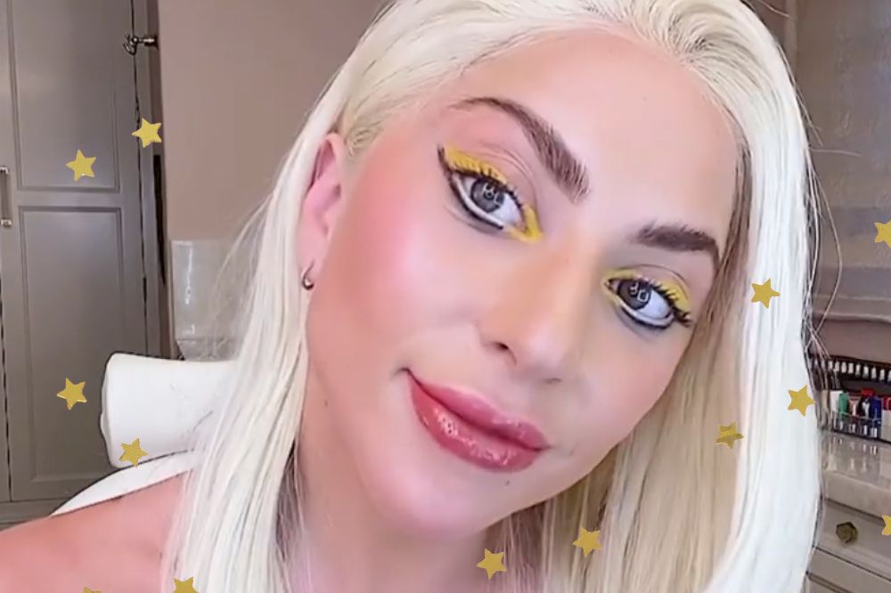 Lady Gaga com a cabeça levemente virada para o lado sorrindo levemente com maquiagem na cor amarela e preta nos olhos; estrelas amarelas decoram a imagem