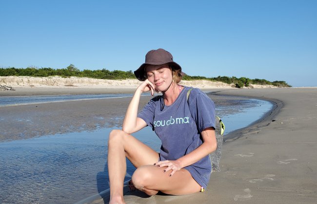 Menina branca de loira, por volta dos 25 anos, descansa sentada na praia com uma camiseta azul com os dizeres 