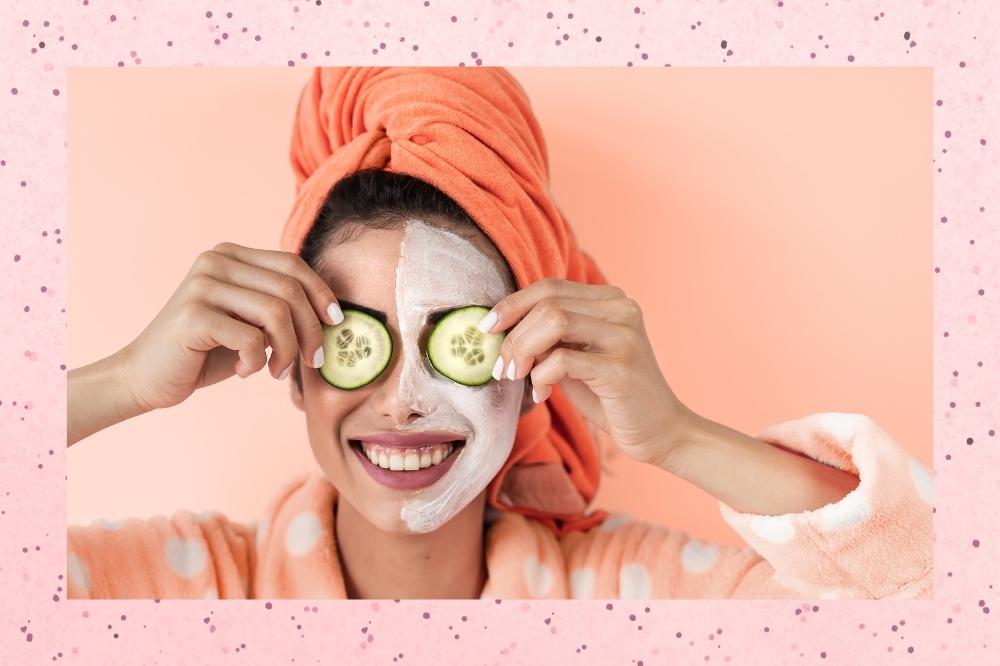 Montagem com o fundo rosa com bolinhas pretas com a foto de uma mulher com toalha na cabeça, roupão e máscara para a pele no rosto no centro.