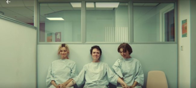 Três mulheres sentadas na sala de espera de uma clínica de aborto. Elas estão de mãos dadas e sorrindo de alívio