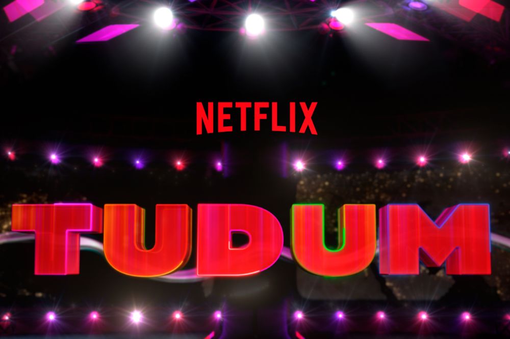 Logo do Tudum Netflix 2022 e vermelho com luzes roxas, rosas e brancas ao fundo