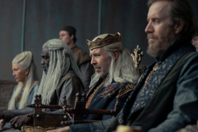 Homens sentados em mesa ao lado do rei, com coroa dourada