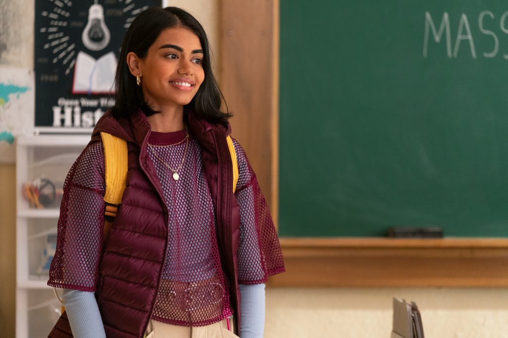 Megan Suri como Aneesa em Eu Nunca...; ela está entrando na sala da aula sorrindo e usando uma mochila amarela com roupas em tons de vinho, azul e roxo