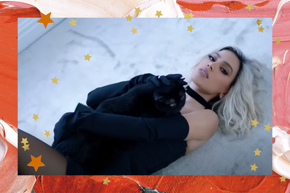 Montagem em fundo vermelho e rosa com estrelinhas douradas de foto de Anitta no clipe da música "Gata" deitada no chão com look preto e segurando um gatinho preto