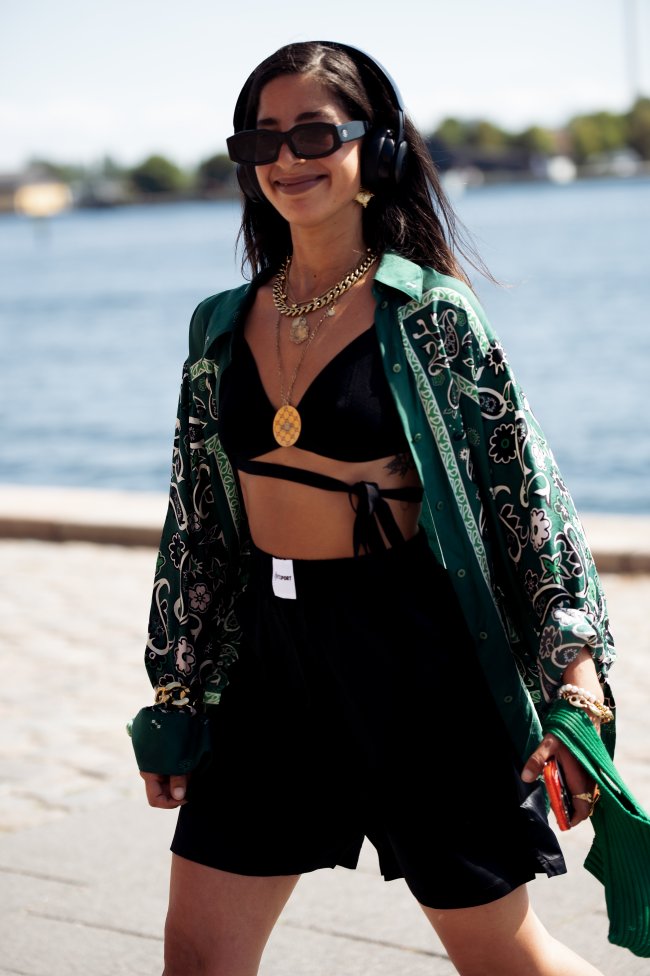 Foto da Selma Kaci na Copenhagen Fashion Week. Na foto ela usa um top cropped preto, calça preto e casaco estampado. Ela está de cabelo solto, usa um fone de ouvido, olha e sorri para a foto.