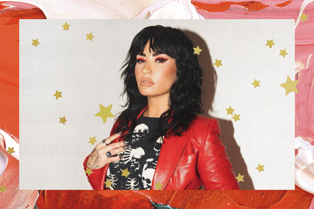 Montagem em fundo vermelho e rosa com estrelinhas douradas de Demi Lovato com expressão facial séria usando maquiagem preta e vermelha, camiseta preta de caveira e jaqueta vermelha por cima