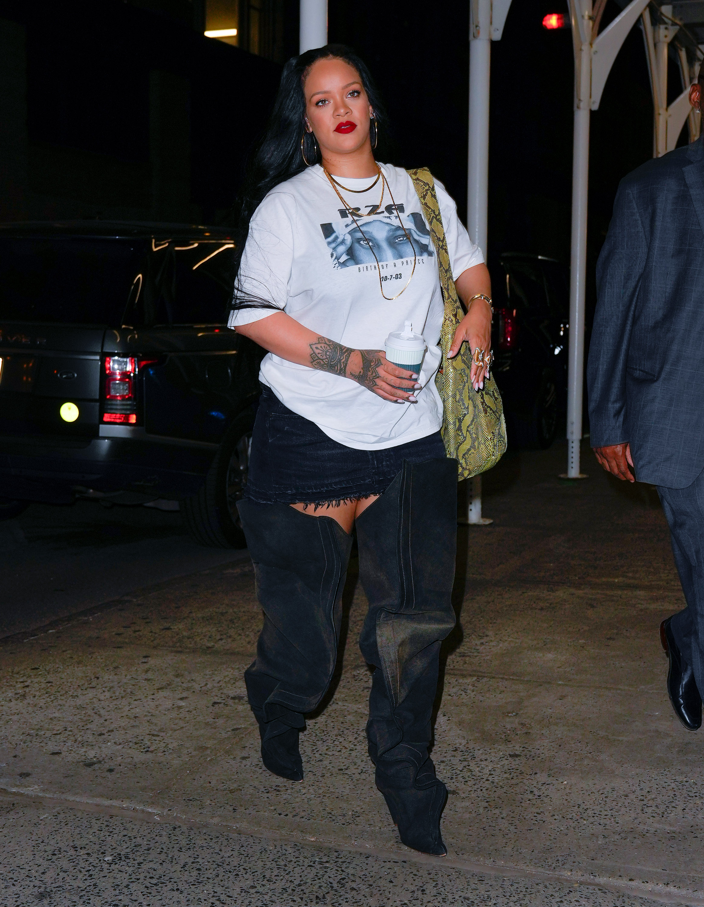 Rihanna usando camiseta branca oversized, minissaia preta e bota slouchy preta que vai até a coxa, além de bolsa com estampa de cobra pendurada no ombro, batom vermelho e brincos de argola